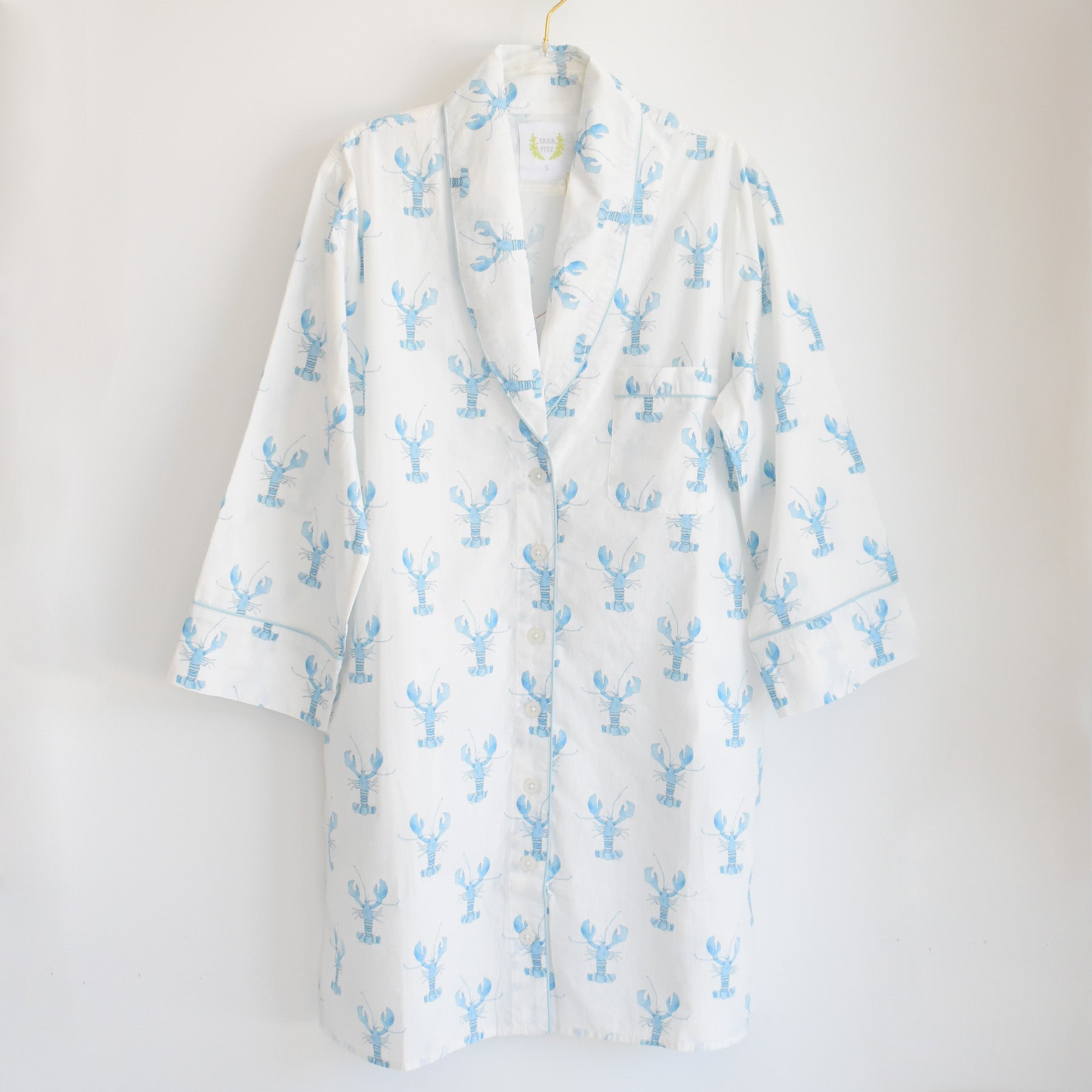 The Blue Lobster Sleep Shirt by Sara Fitz, featuring long white cotton sleeves and a playful blue lobster print, hangs on a gold hanger against a plain white background—bringing the perfect touch of whimsy to women’s sleepwear.