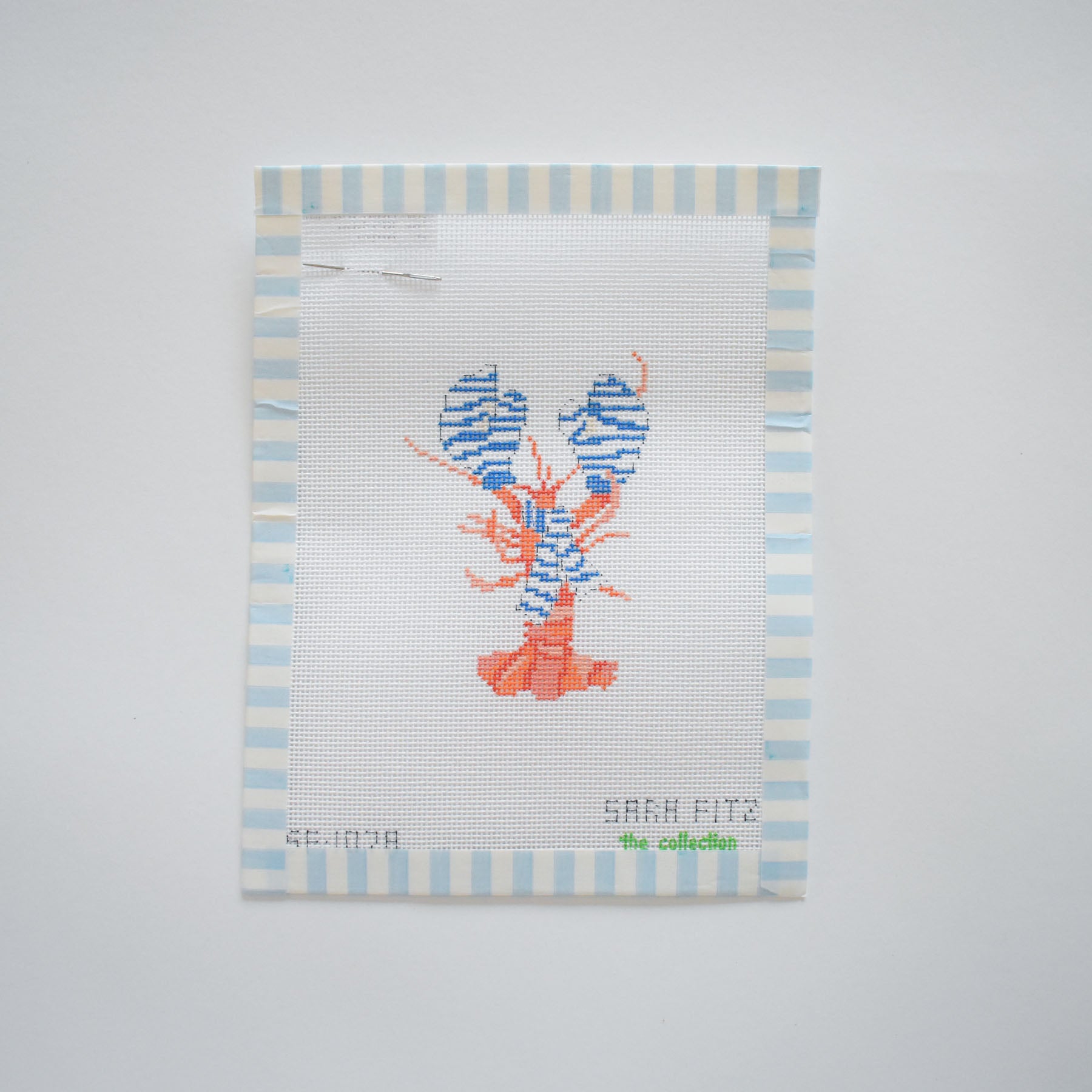 The Small Cozy Lobster Needlepoint by Sara Fitz features an orange and blue striped lobster on white canvas with light blue and white borders. “SARAH FITZ the collection” is stitched at the bottom. 18 mesh canvas only.