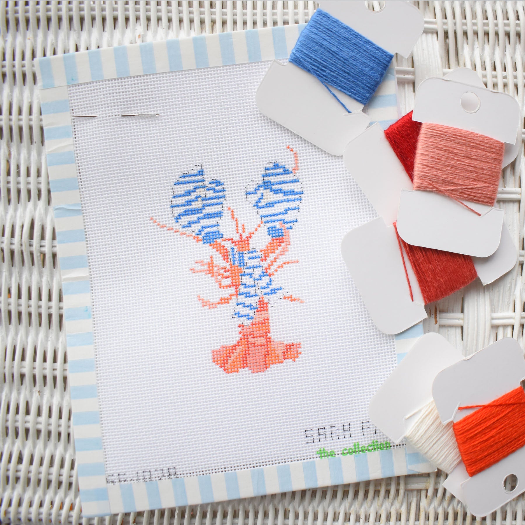 The Small Cozy Lobster Needlepoint by Sara Fitz features a partially stitched blue and orange lobster on white canvas, with blue, red, orange, and white threads—ideal for 18 mesh canvas fans.