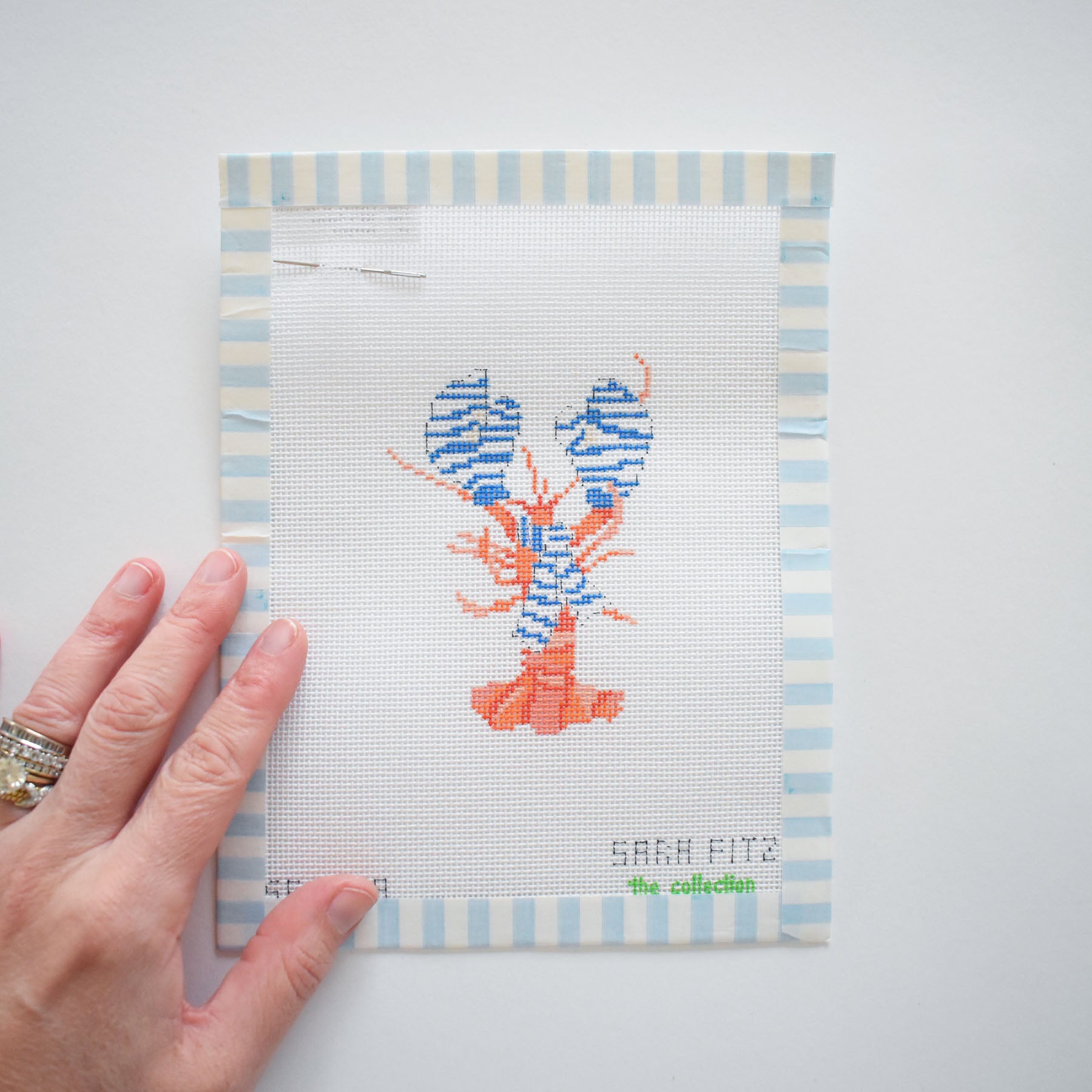 A hand with rings holds the Small Cozy Lobster Needlepoint by Sara Fitz—an 18 mesh canvas with blue and white striped border, featuring a cross-stitched blue and orange lobster and "SARAH FITZ" stitched at the bottom right.