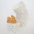 A stack of Sara Fitz Hermit Crab Snack Cups, featuring colorful seashell patterns, with an 8.5 oz cup in front holding a large cookie and crumbs on a white surface.