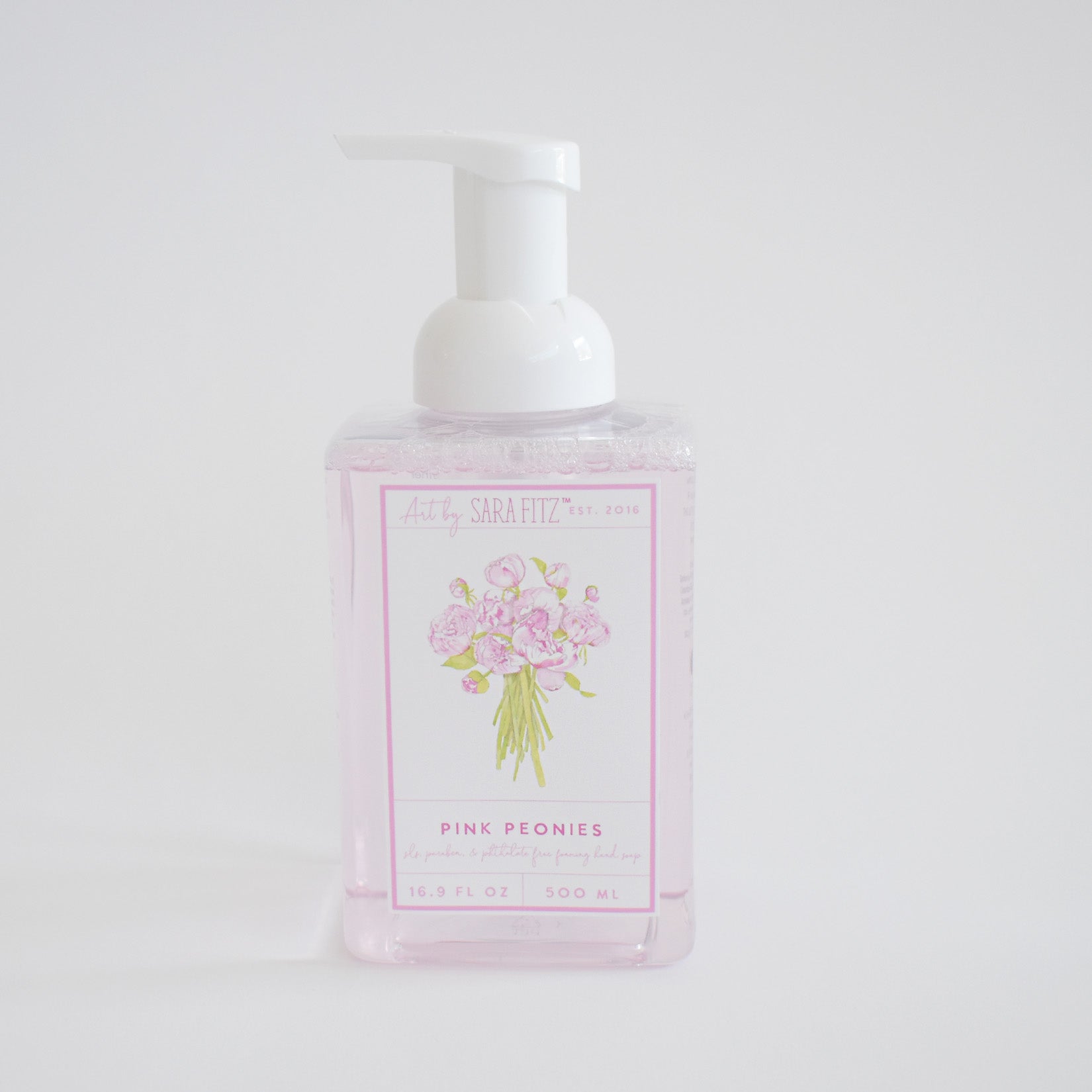 A clear pump bottle of Sara Fitz Foaming Soap Peony, filled with light pink, non-toxic liquid and adorned with a floral illustration, is displayed against a plain white background.