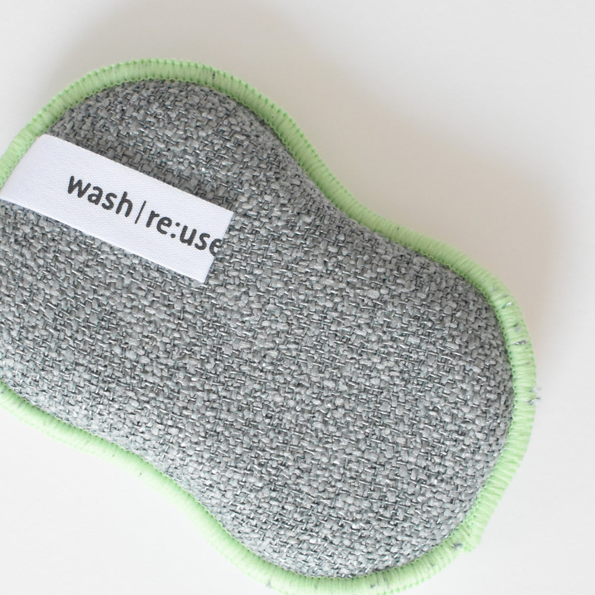 A gray, textured, mitten-shaped Re:usable Sponge, Mittens by Sara Fitz with a light green stitched edge and a white label that reads wash|re:use.