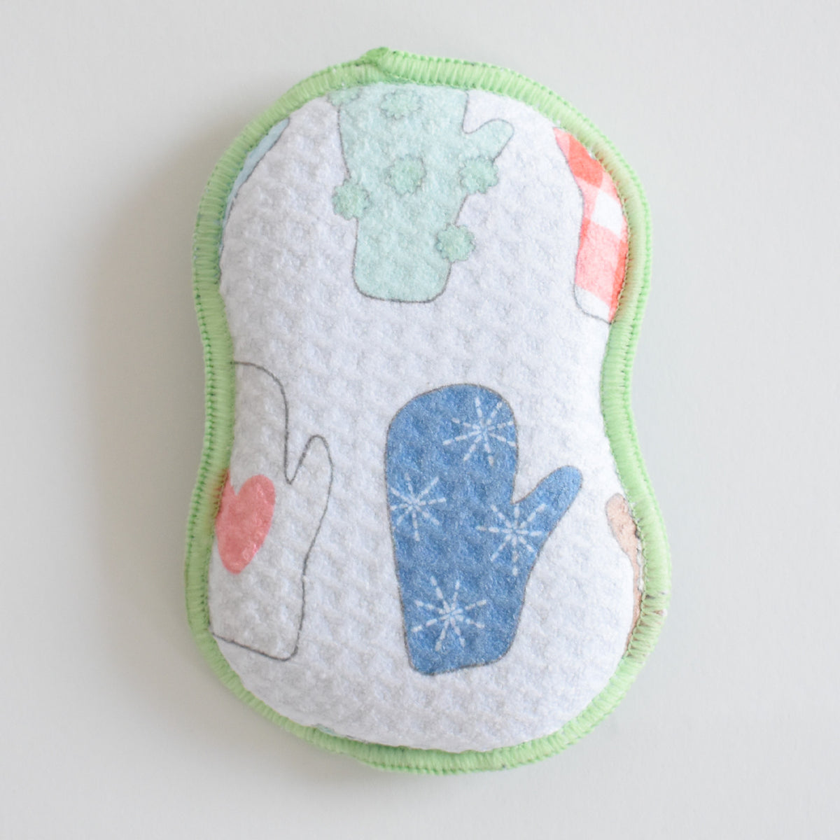 An hourglass-shaped Re:usable Sponge, Mittens with a light green border, featuring a white surface patterned with blue, green, and red mittens.