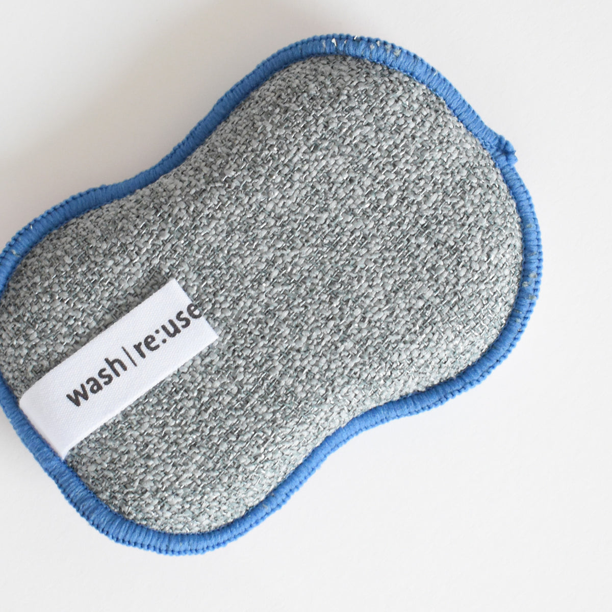 A textured gray Re:usable Sponge, Ornament with blue trim and a small tag that reads wash|re:use.
