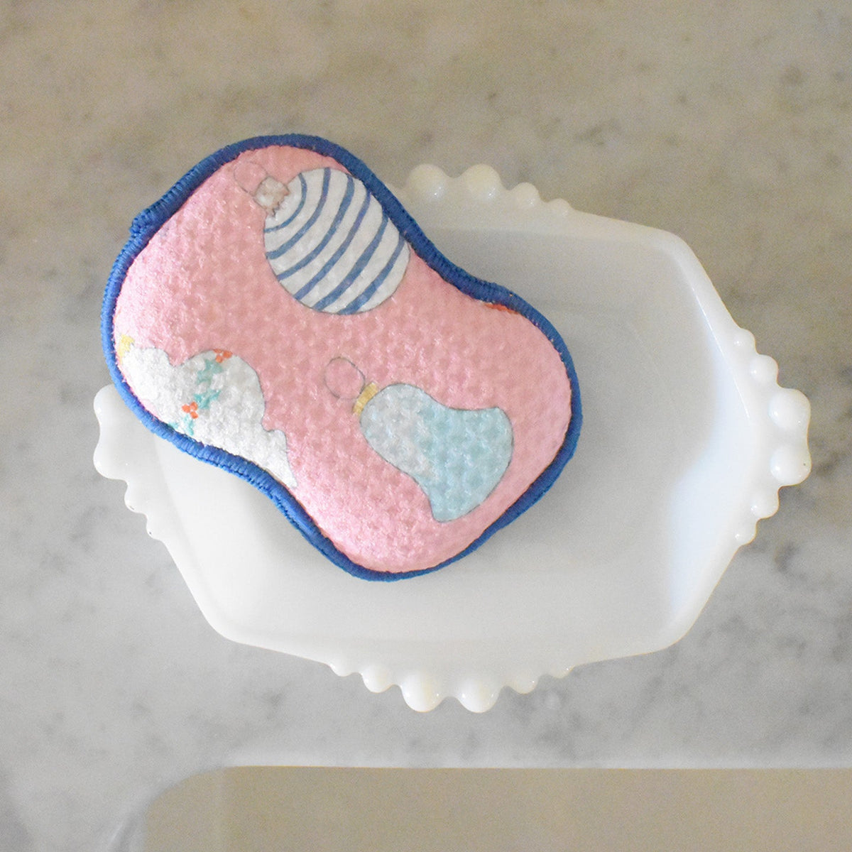 Sara Fitz Re:usable Sponge, Ornament, featuring a pink sponge with blue trim and a pattern of holiday ornaments sitting in a white dish.