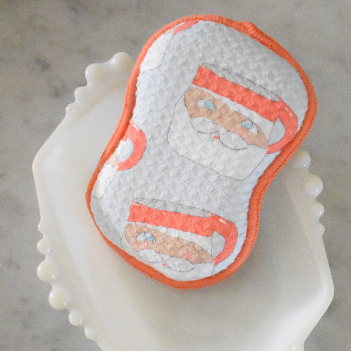 A Re:usable Sponge, Santa Mug with a repeating Santa Claus face pattern and an orange stitched edge, resting in a white scalloped dish for eco-friendly cleaning.