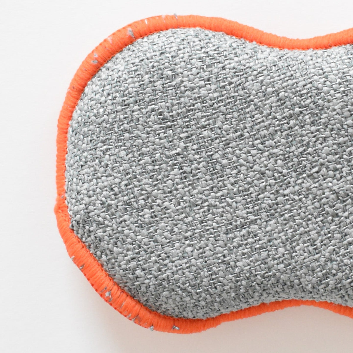 A grey, textured, reusable Sara Fitz Re:usable Sponge with orange stitched edges.