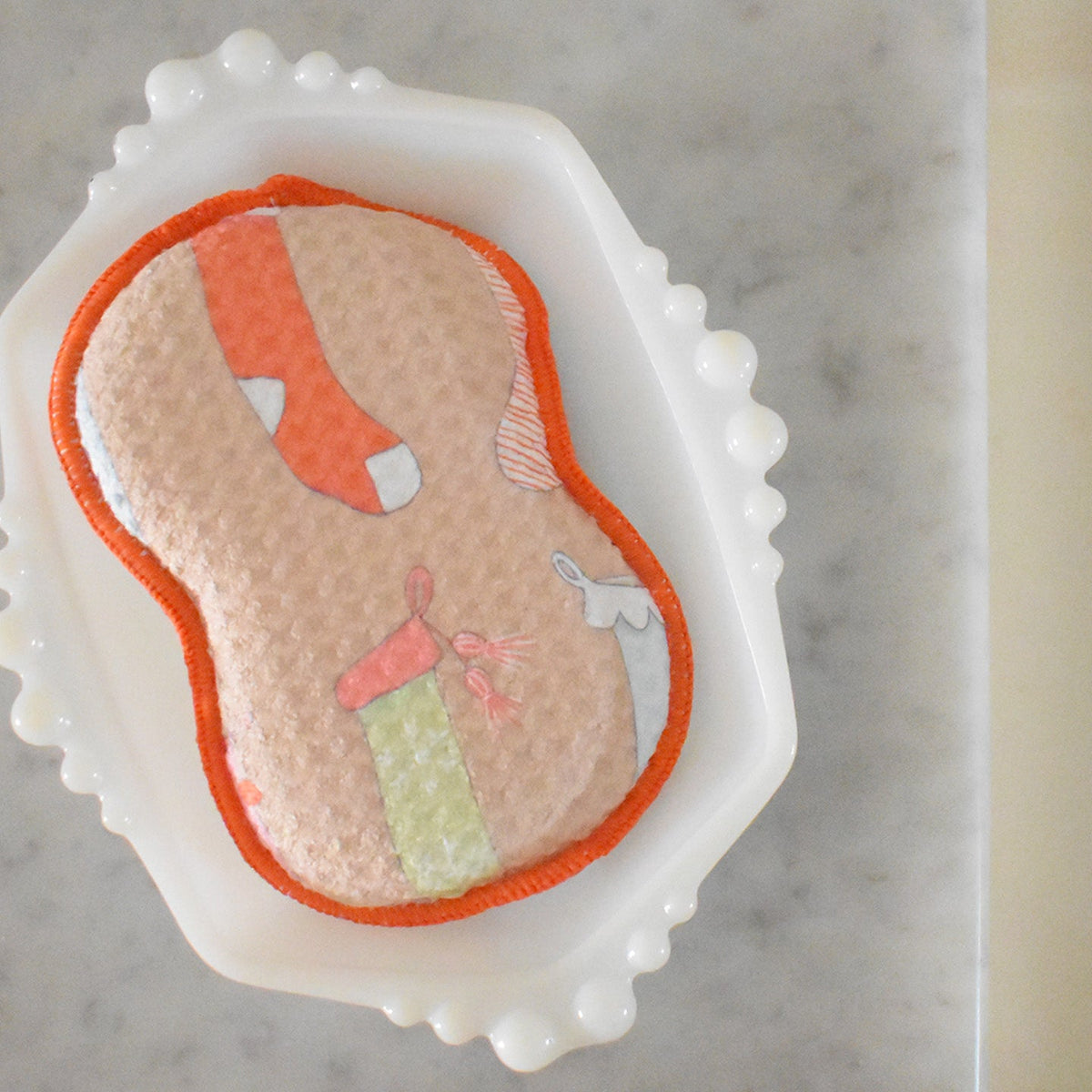 A textured, hourglass-shaped Sara Fitz Re:usable Sponge, Stockings, with an orange border and a pattern of stockings, resting on a white milk glass dish.