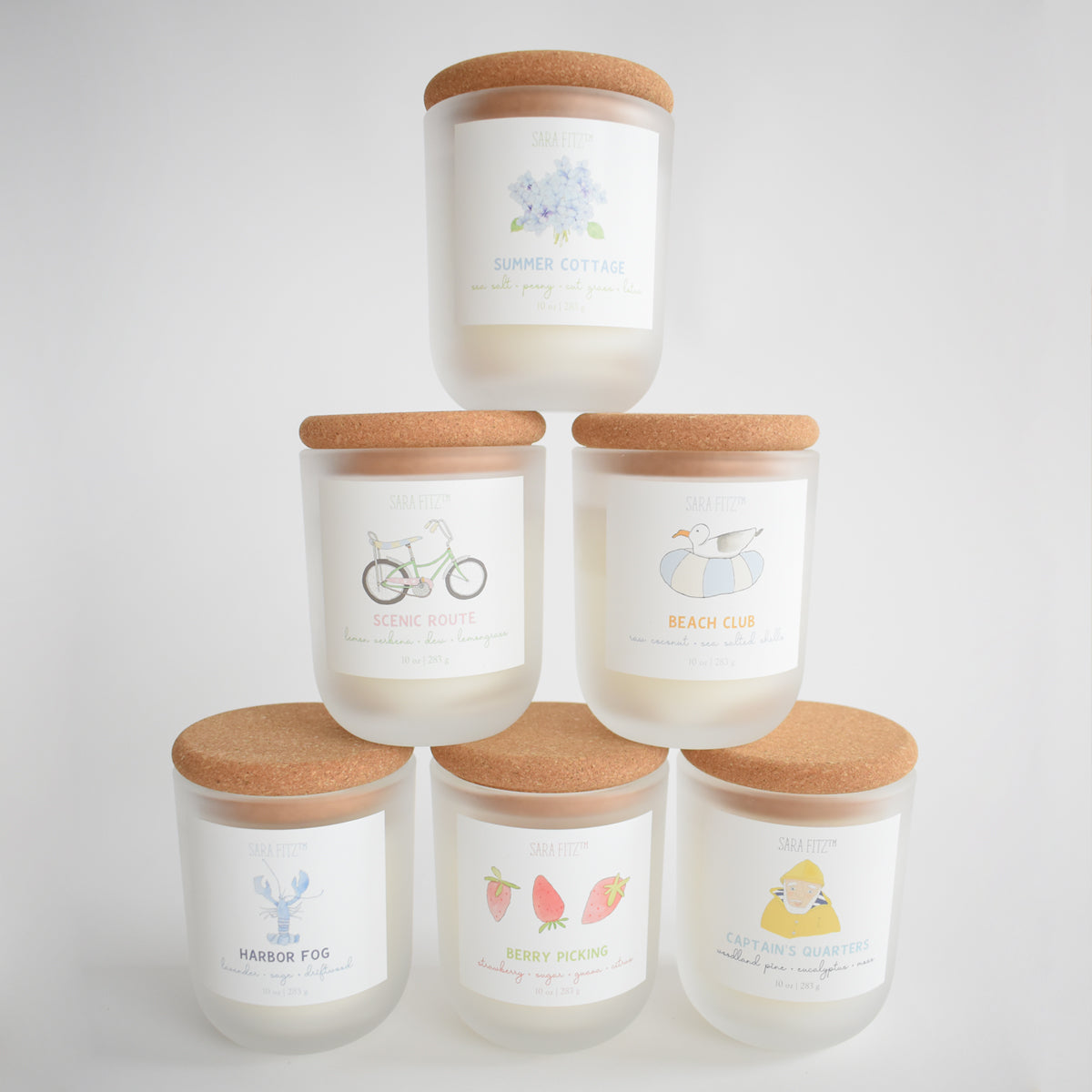 Six white Summer Cottage candles by Sara Fitz, hand-poured in Maine with coconut soy wax, are stacked in a pyramid. Each features a pastel illustration and names such as Summer Cottage, Scenic Route, Harbor Fog, and Captain’s Quarters.