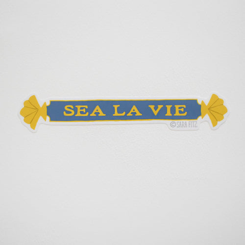 The Sara Fitz Quarterboard Sticker features shell designs at each end, bold yellow "SEA LA VIE" text on blue with yellow edges. Weatherproof and made in the USA, it stands out against a white wall.