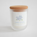 The Sara Fitz Summer Cottage Candle is hand-poured in Maine in a frosted glass jar with cork lid. The label features a watercolor hydrangea. Made with coconut soy wax, it has scent notes of sea salt, peony, cut grass, and lilac.