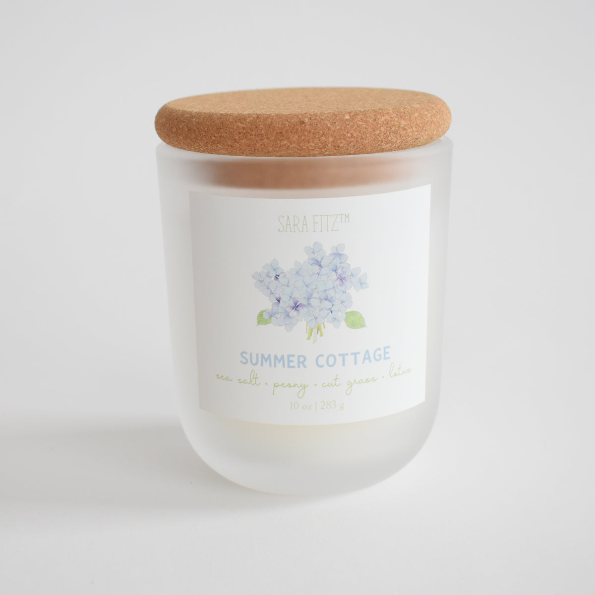 The Sara Fitz Summer Cottage Candle is hand-poured in Maine in a frosted glass jar with cork lid. The label features a watercolor hydrangea. Made with coconut soy wax, it has scent notes of sea salt, peony, cut grass, and lilac.