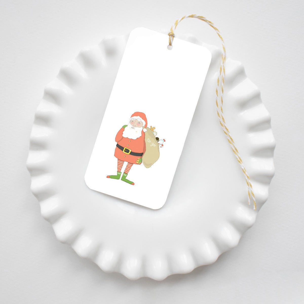 Santa Fitz Tags featuring an illustration of Santa Claus carrying a sack, attached to yellow and white baker’s twine and resting on a white scalloped dish.
