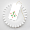 A rectangular Holly Cross Stitch Tags gift tag featuring a red and green cross-stitched holly pattern, tied with yellow and white baker’s twine and resting on a white ruffled plate.