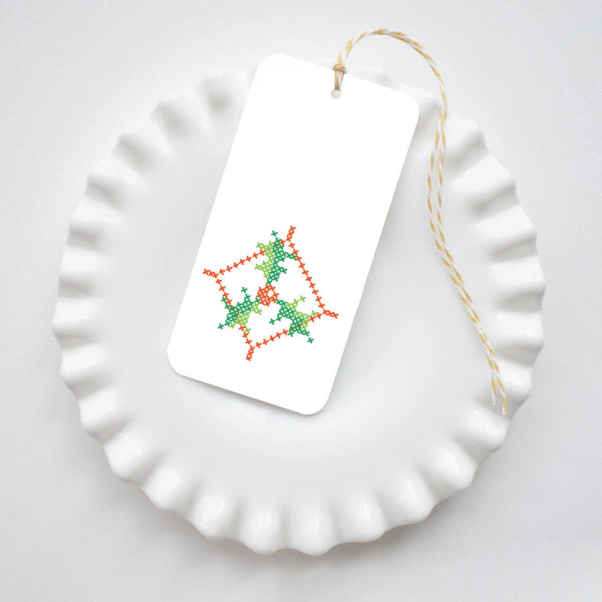 A rectangular Holly Cross Stitch Tags gift tag featuring a red and green cross-stitched holly pattern, tied with yellow and white baker’s twine and resting on a white ruffled plate.