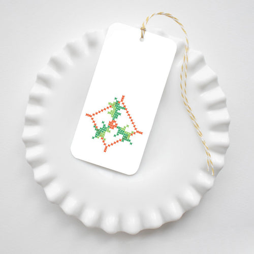 A rectangular Holly Cross Stitch Tags gift tag featuring a red and green cross-stitched holly pattern, tied with yellow and white baker’s twine and resting on a white ruffled plate.