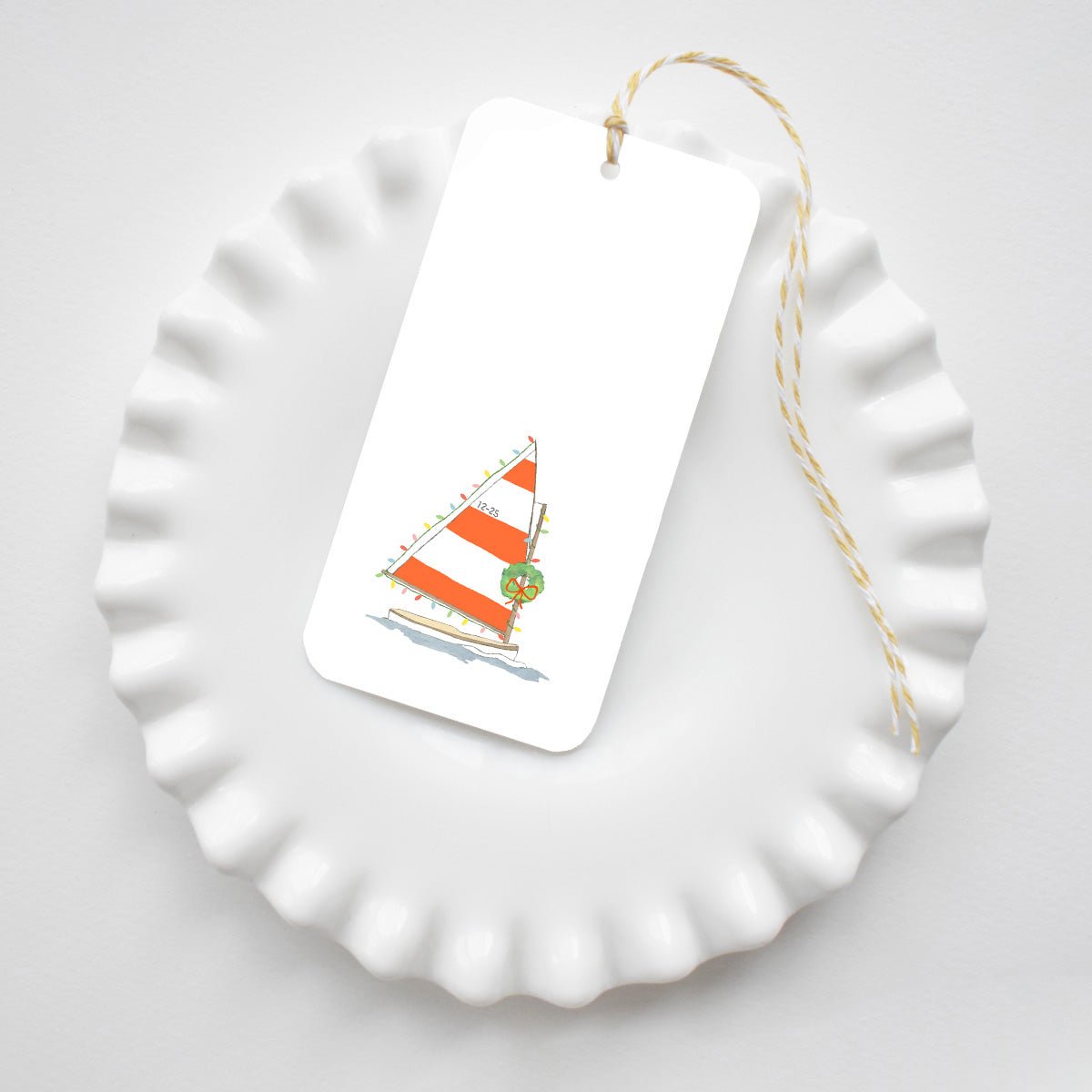 Holiday Sailboat Tags featuring an illustration of a sailboat with red and white striped sails decorated with Christmas lights and a wreath, hanging on yellow and white baker’s twine.