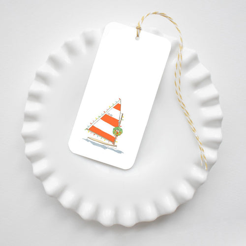 Holiday Sailboat Tags featuring an illustration of a sailboat with red and white striped sails decorated with Christmas lights and a wreath, hanging on yellow and white baker’s twine.