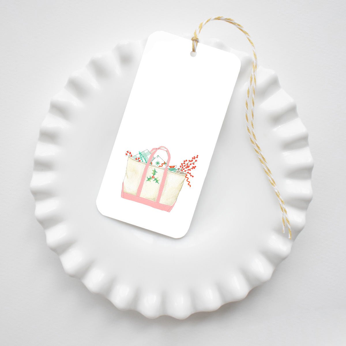 Sara Fitz Holiday Tote Tags: a rectangular gift tag featuring a watercolor illustration of a tote bag filled with holiday greenery, resting on a white fluted plate tied with yellow and white baker’s twine.