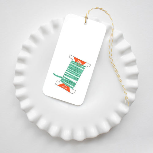 Holiday Trimmings Tags are rectangular gift tags tied with yellow and white baker’s twine, featuring an illustration of a green ribbon spool labeled jolly and 2yd cotton.