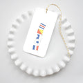 A COAST Flags Tag by Sara Fitz, featuring nautical signal flags and gold-and-white baker's twine, is displayed on a white fluted-edge plate against a plain white background.
