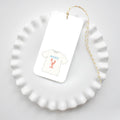 A white ruffled plate holds a Maine Tee Tag by Sara Fitz—a rectangular gift tag with baker’s twine, featuring a T-shirt labeled “MAINE” with a red lobster graphic. A plain white background highlights its playful design.