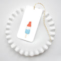 A white scalloped plate holds a rectangular Sara Fitz Popsicle Tag, tied with yellow and white baker’s twine and featuring a red, white, and blue popsicle illustration.