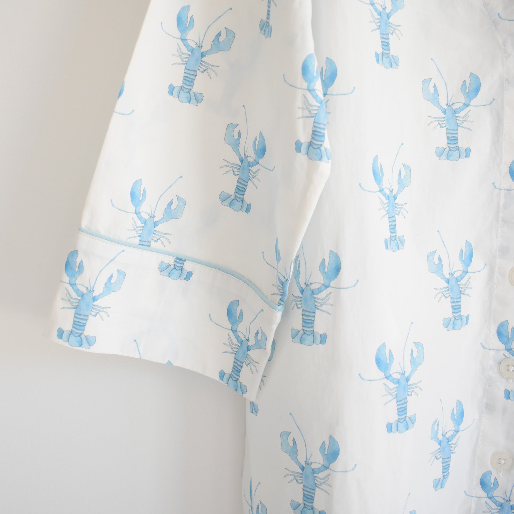 The Sara Fitz Blue Lobster Sleep Shirt features a white fabric adorned with a light blue lobster pattern, shown from the side with short sleeves and partially visible white buttons—a charming women’s sleepwear option that adds playful flair to bedtime style.