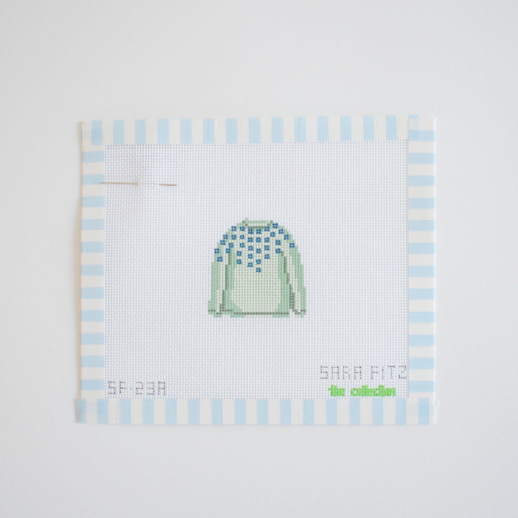 The MINI Pom Sweater Needlepoint by Sara Fitz is shown in progress on 18 mesh white canvas bordered with blue and white striped tape, featuring a green sweater with blue detail. A needle and unfinished stitches appear on the left.