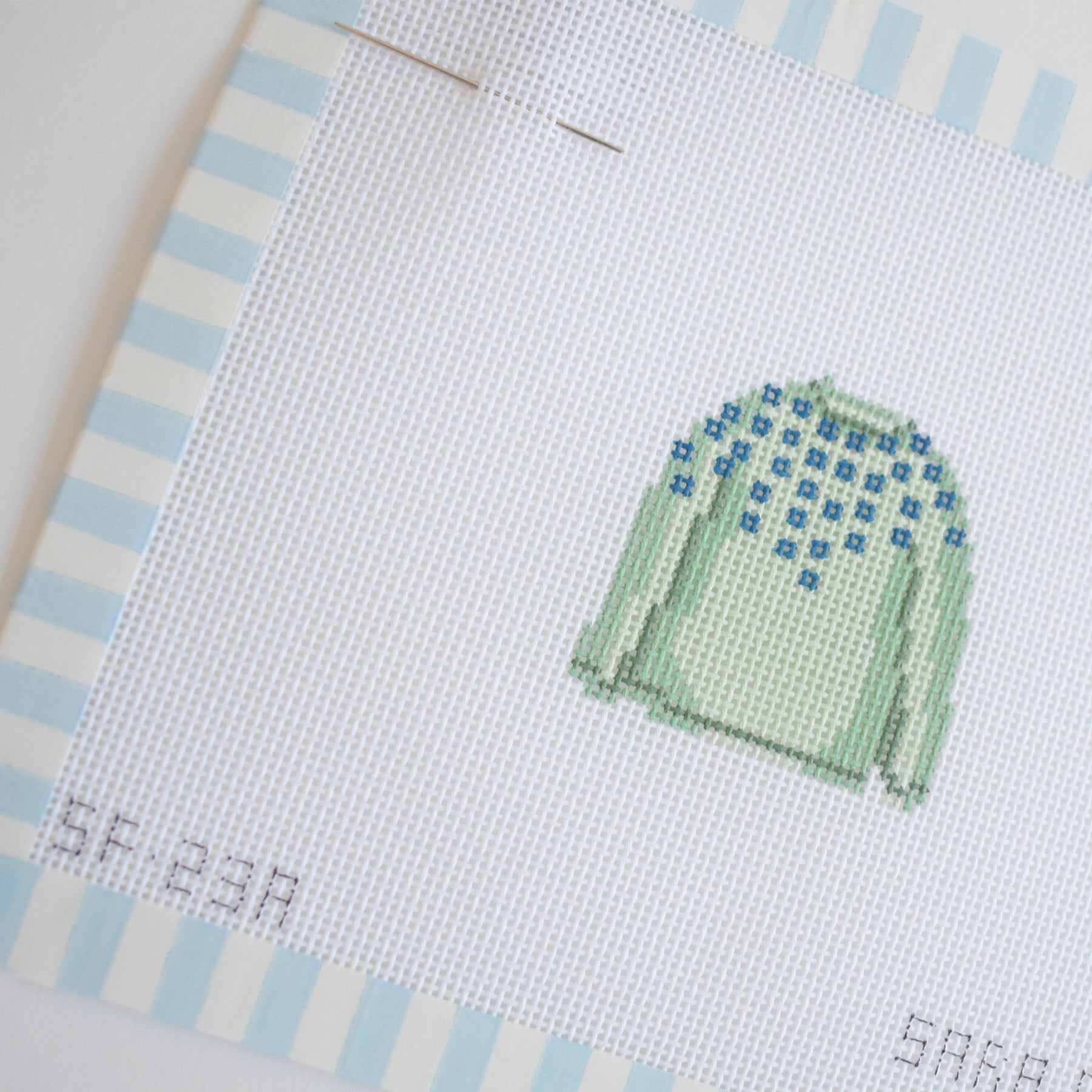 The MINI Pom Sweater Needlepoint by Sara Fitz features a stitch-painted light green sweater with blue patterns on the shoulders, displayed on white canvas with blue and white striped edges and two needles pinned at the top of the 18 mesh canvas.