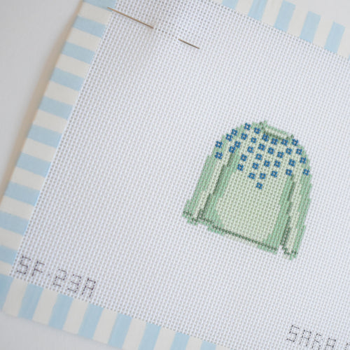 The MINI Pom Sweater Needlepoint by Sara Fitz features a stitch-painted light green sweater with blue patterns on the shoulders, displayed on white canvas with blue and white striped edges and two needles pinned at the top of the 18 mesh canvas.