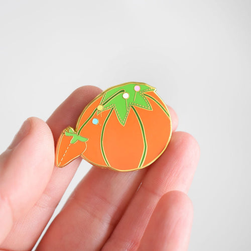 A hand holds the Sara Fitz Tomato Pincushion Needle Minder—an enamel pin shaped like an orange tomato with a green stem and leaves, plus a small strawberry detail. Perfect as an enamel needle minder or stitching accessory, this charming piece stands out beautifully against the plain, light gray background.