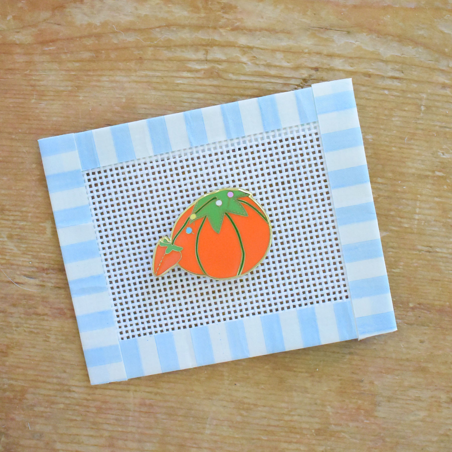 The Sara Fitz Tomato Pincushion Needle Minder features a charming small orange tomato-shaped pin cushion illustration on white plastic mesh, bordered by blue and white striped tape—a delightful stitching accessory ideal for organizing your needles or pairing with enamel needle minders.