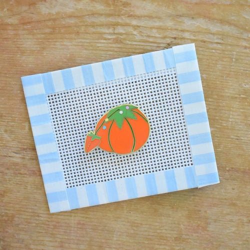 The Sara Fitz Tomato Pincushion Needle Minder features a charming small orange tomato-shaped pin cushion illustration on white plastic mesh, bordered by blue and white striped tape—a delightful stitching accessory ideal for organizing your needles or pairing with enamel needle minders.