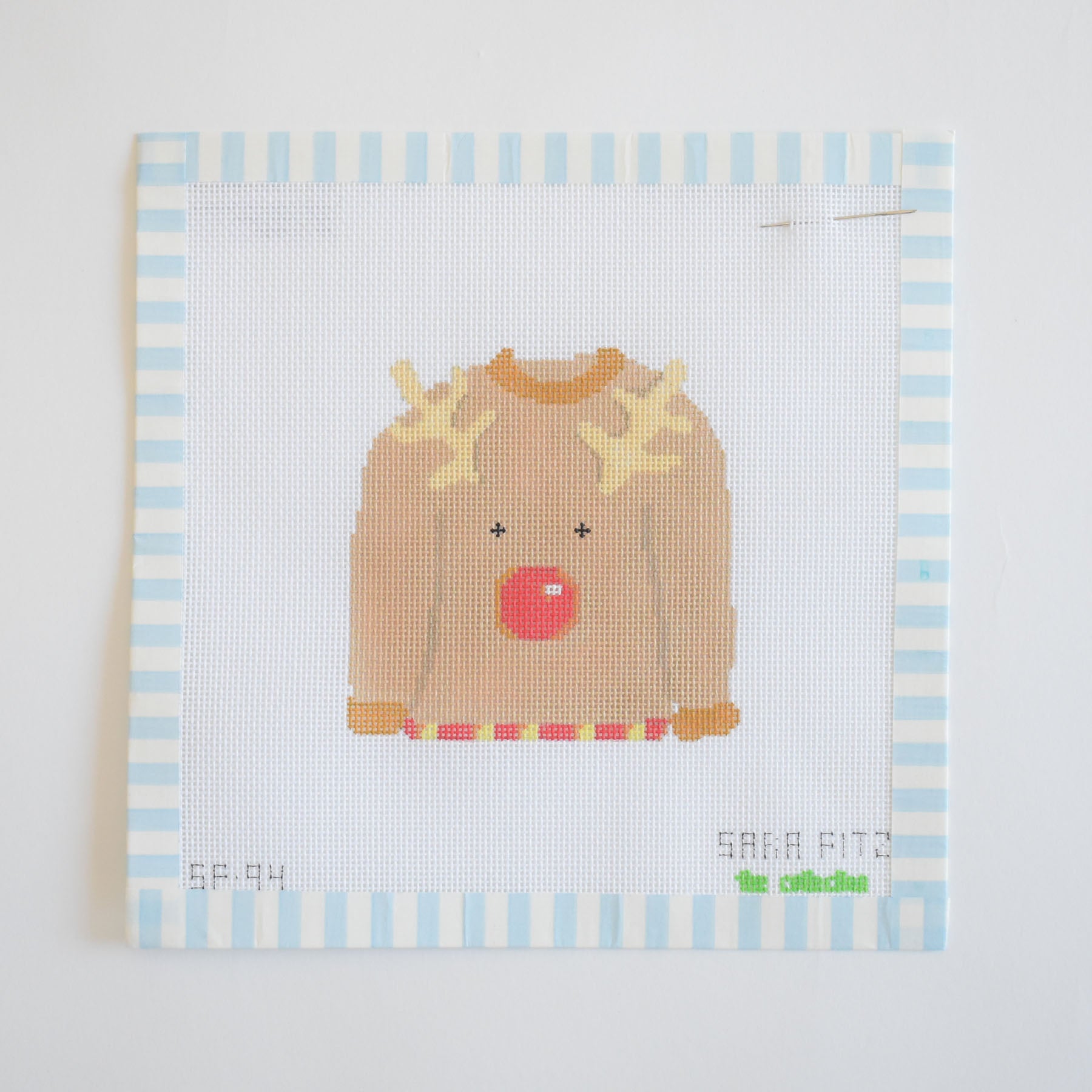The Sara Fitz Ugly Sweater Needlepoint is a stitch-painted 18 mesh canvas featuring a brown reindeer sweater with a red nose and yellow antlers on a blue and white striped border.