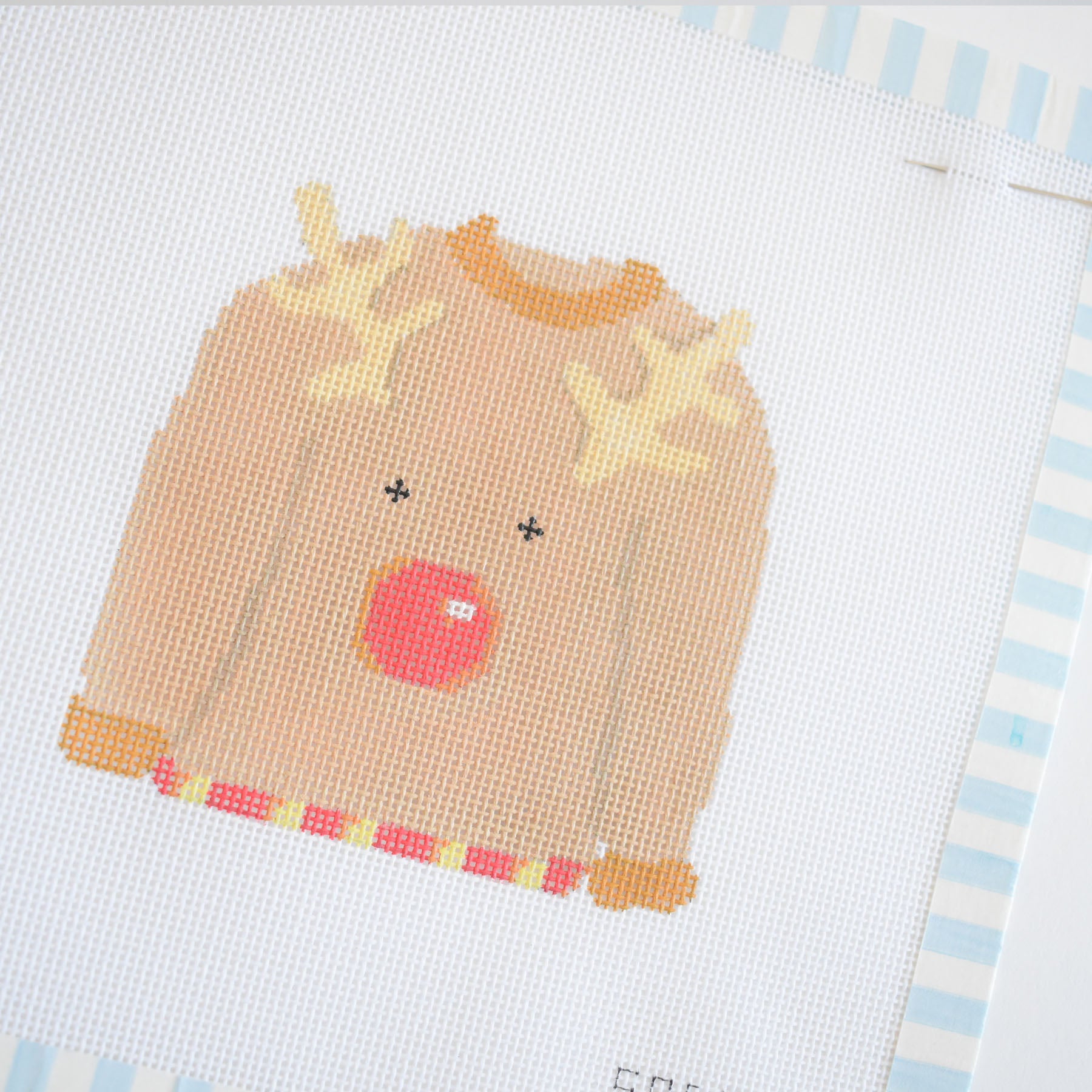 The Sara Fitz Ugly Sweater Needlepoint features a light brown sweater with a reindeer face—antlers, black eyes, and red nose—on white fabric with a light blue striped border, designed for an 18 mesh canvas.