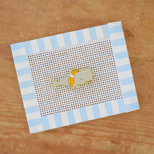 The Sara Fitz Whale Needle Minder features a small gray whale with an orange patch, centered on white mesh canvas with a blue and white striped frame—an ideal stitching accessory for your projects.