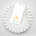 A white scalloped-edged plate displays a Block Print Sherbet Tag by Sara Fitz, featuring a yellow flower illustration and yellow baker’s twine, set on a white background.