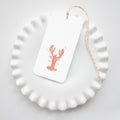 A white scalloped-edge plate displays a Sara Fitz Lobster Tag—a white gift tag with a red lobster illustration and yellow-and-white baker's twine—carefully arranged on top.