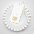 A white plate displays a Nantucket Basket Tag by Sara Fitz, featuring a yellow woven basket illustration and yellow-and-white baker's twine, all shown against a white background.