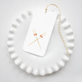 The Oar Tags by Sara Fitz feature two crossed oars on a white gift tag, attached with yellow and white baker’s twine. Shown on a ruffled-edge ceramic plate, these tags are perfect for any occasion and complement an all-white setting.