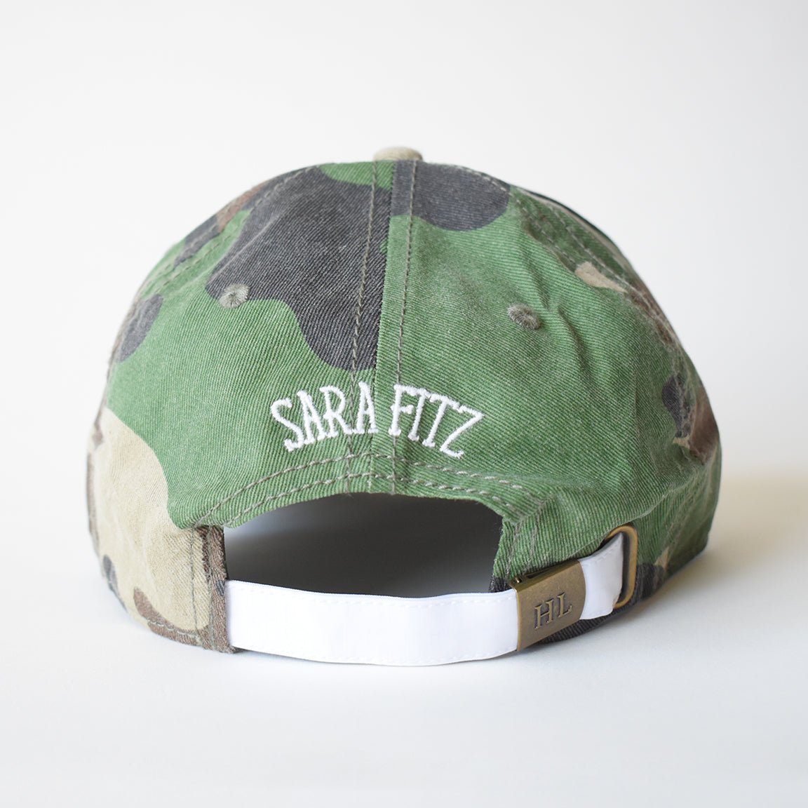 The Striped Shirt Hat, Camo by Sara Fitz is a cotton baseball cap with a camouflage pattern, white SARA FITZ needlepoint above the opening, and an adjustable back strap featuring a metal clasp engraved with HIL.