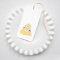 A white, scalloped-edge plate holds a Sara Fitz Skipper Tag—a rectangular gift tag featuring an elderly person in a yellow raincoat and hat—tied with yellow and white baker’s twine on top.