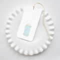A white ruffled-edge plate displays a 2x4" Sara Fitz Thermos Tag, featuring a blue thermos illustration and finished with yellow-and-white baker’s twine.